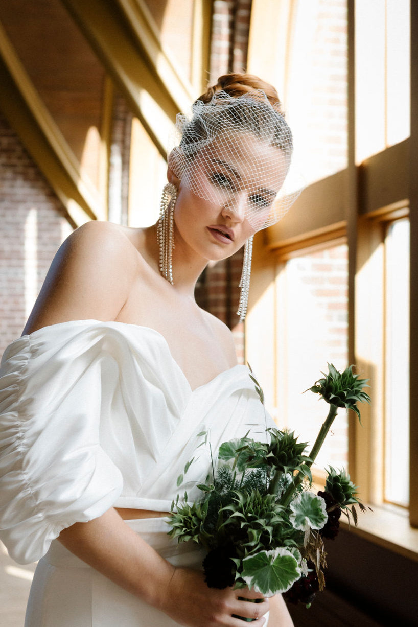 a woman wearing an off the shoulder wedding dress is standing in front of window holding a large green floral bouquet.  The woman is wearing a vintage birdcage veil and long dangling earrings