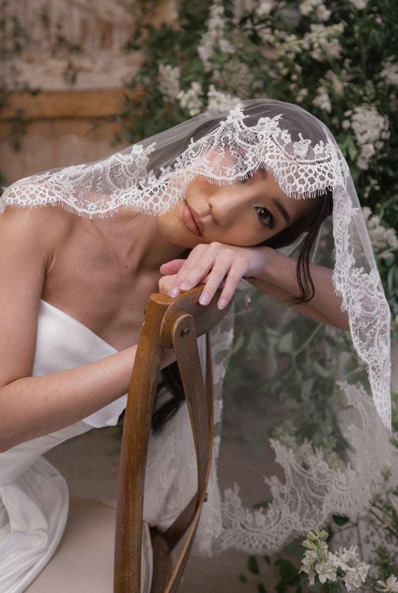 a woman is wearing a white wedding dress and learing on the back of a chair.  She has a lace trimmed white veil on with green foliage in the background