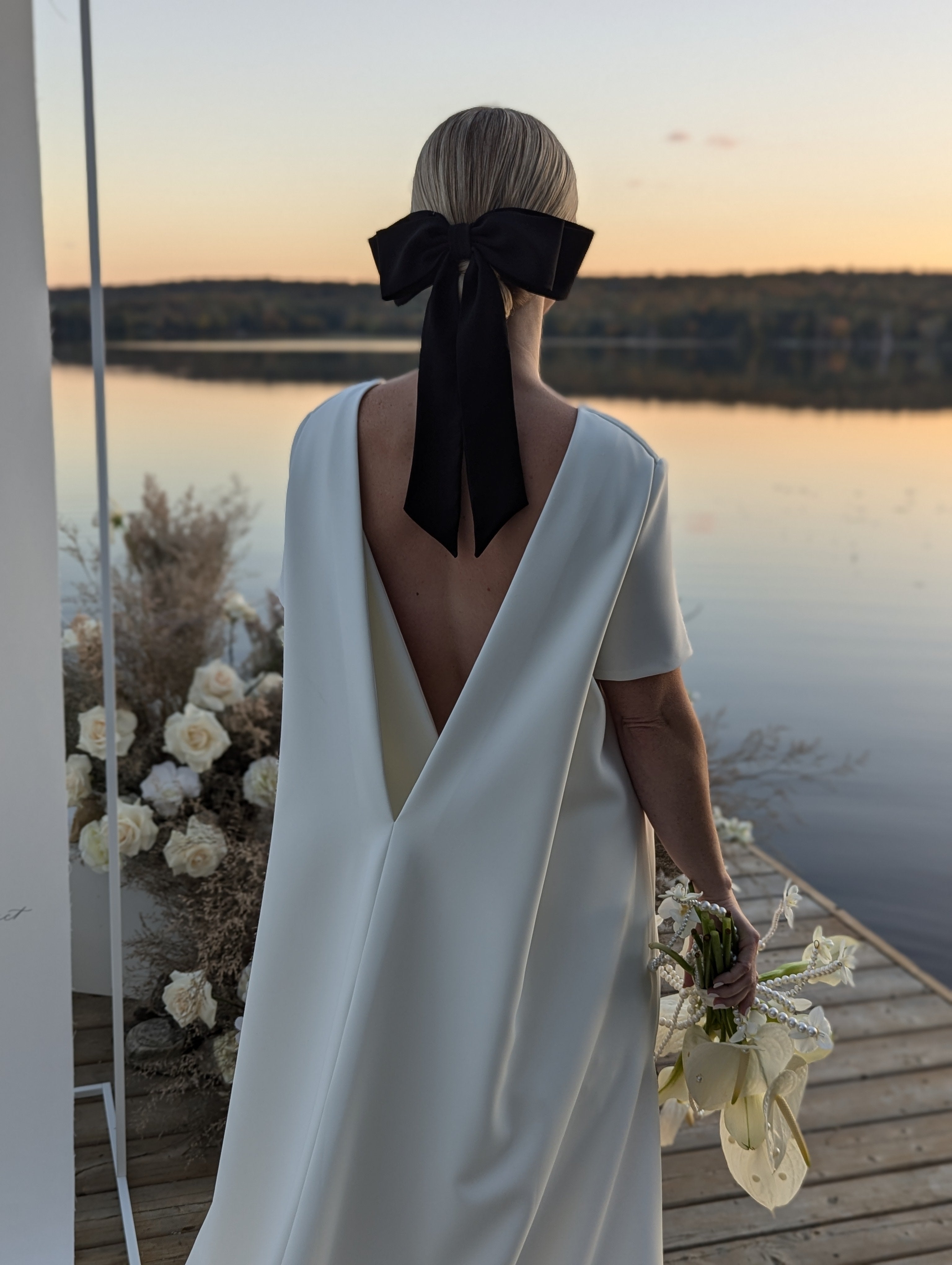 a woman is standing in front of the lake on a dock wearing a modern open back wedding dress and holding a white bouquet.  She is wearing a large black bow in her hair.