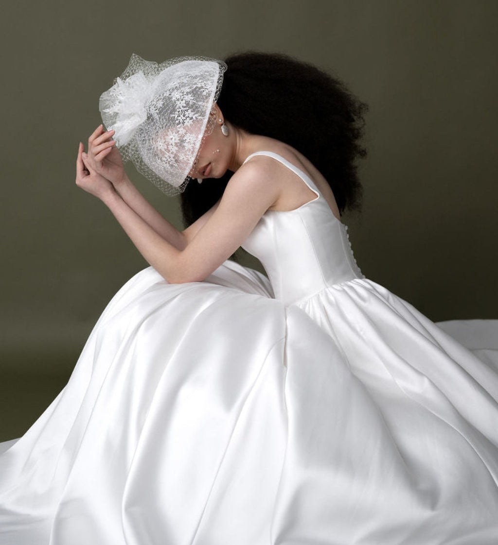bride in a white dress is wearing a large lace hat with pearl earrings against an olive green backdrop
