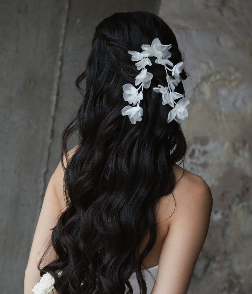 a bride is standing infront of a textured background with long black wavy hair and has cascading silk flowers in her hair