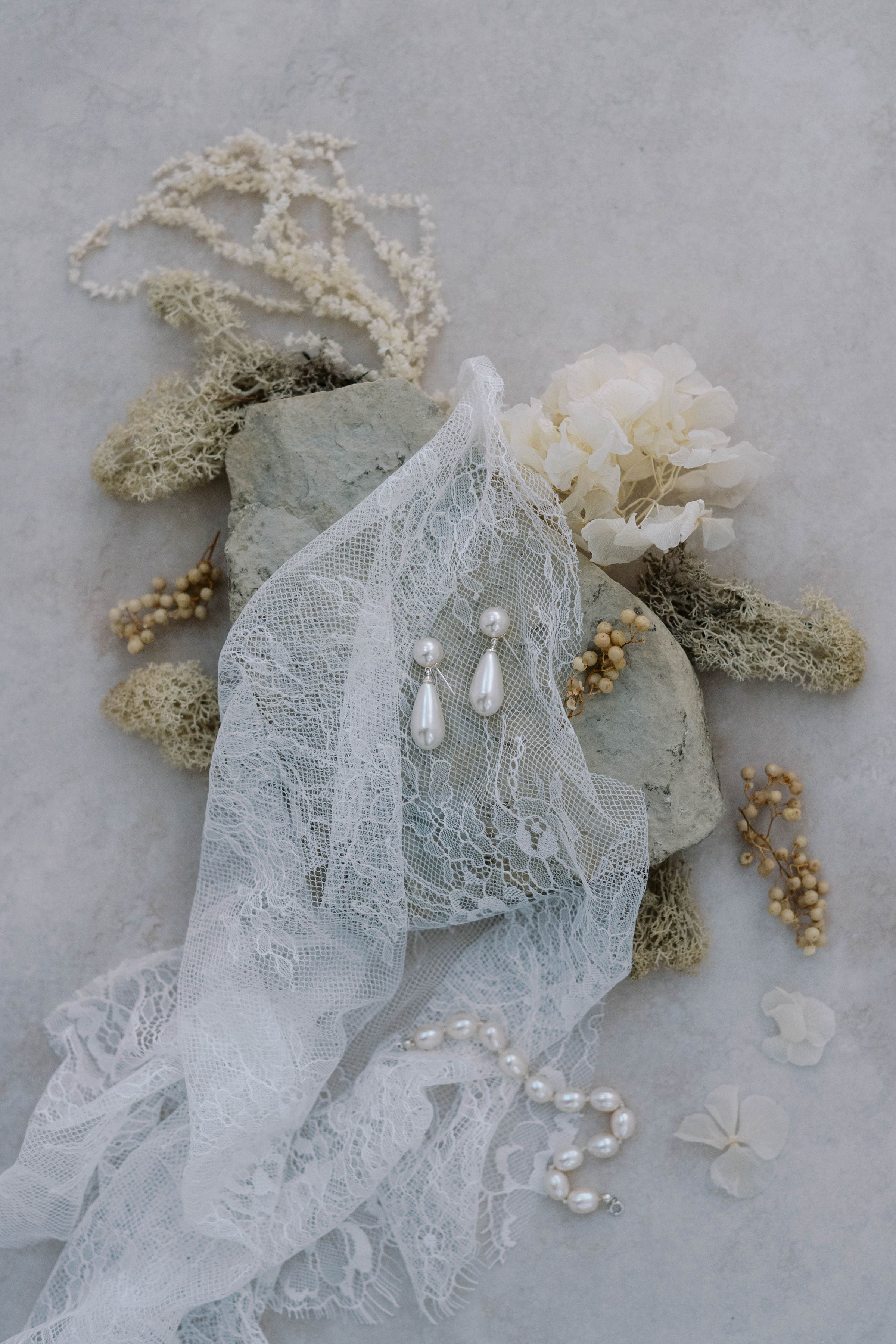  lace is laid overtop of a textured stone accented with dried florals.  pearl earrings and a pearl bracelet are displayed ontop of the lace and stone