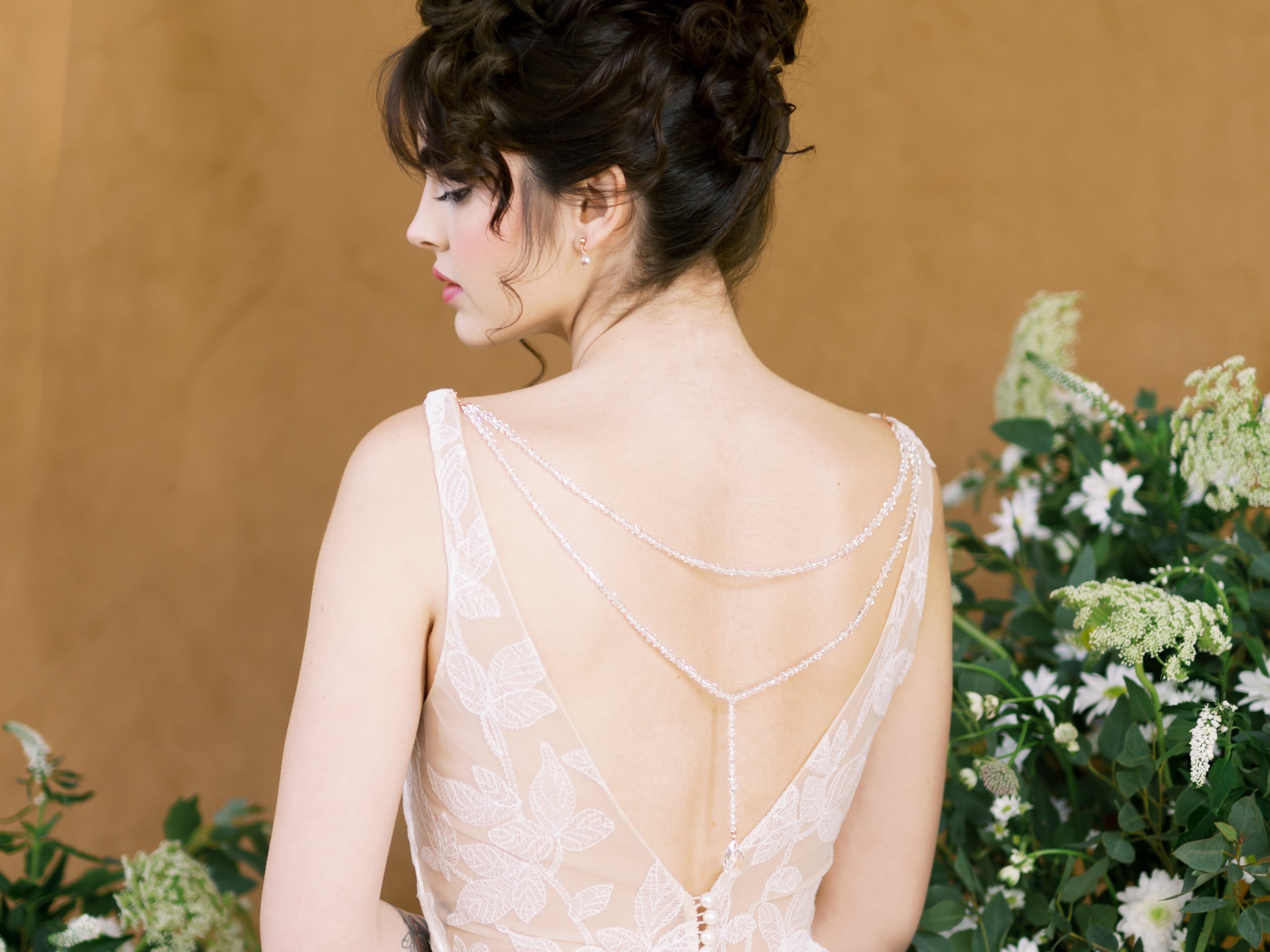  a woman is sittingn amongst green and white florals is wearing a low back lace wedding dress and is wearing a crystal beaded back necklace that attaches to her wedding dress