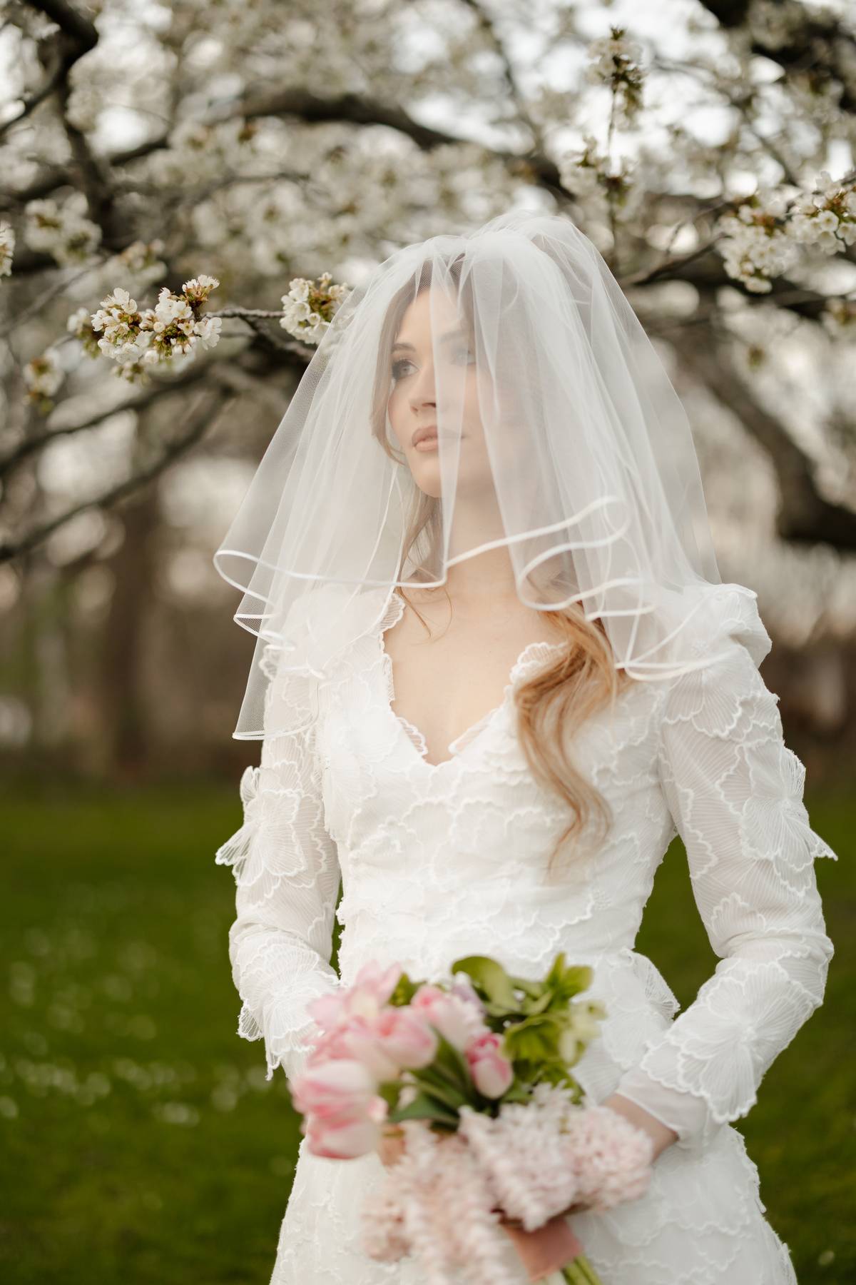 A bride in a white lace wedding dress with a sheer veil holds a bouquet of pink flowers outdoors in front of blooming blossom trees.