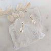 Pair of gold and pearl drop earrings on a textured white surface with dried leaves.