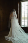 Person wearing a white lace wedding dress with a long train in a room with wooden walls and a window.