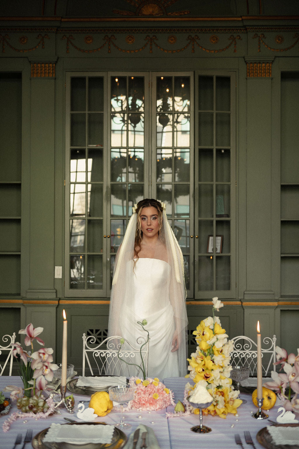 A bride in a strapless white wedding gown and long veil stands centered in front of a large green-paneled window. A decorated dining table with yellow flowers, candles, plates, and lemon slices is visible in the foreground.