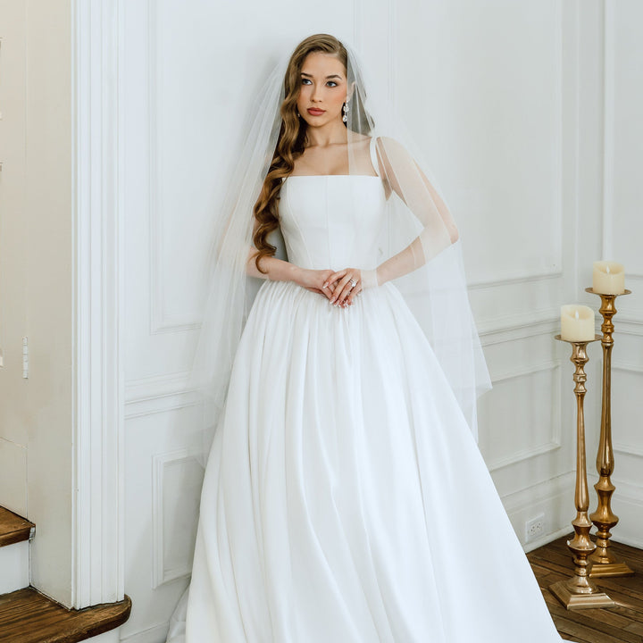 Woman in a white wedding dress with a veil standing in a room with white walls and wooden floor.