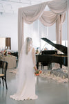 Woman in a white dress holding flowers in a room with a grand piano and draped fabric.
