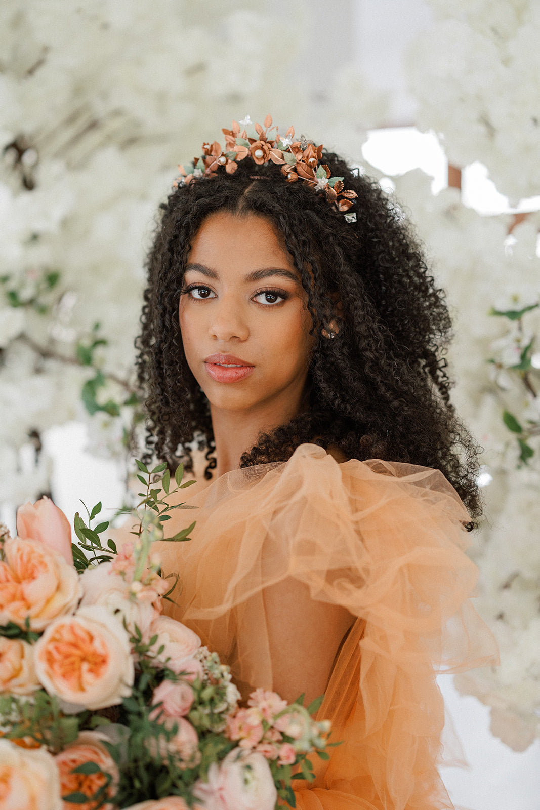 Woman in an orange dress with a floral headpiece and bouquet, surrounded by white flowers.