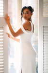 Woman in a white dress standing in front of a window with blinds.
