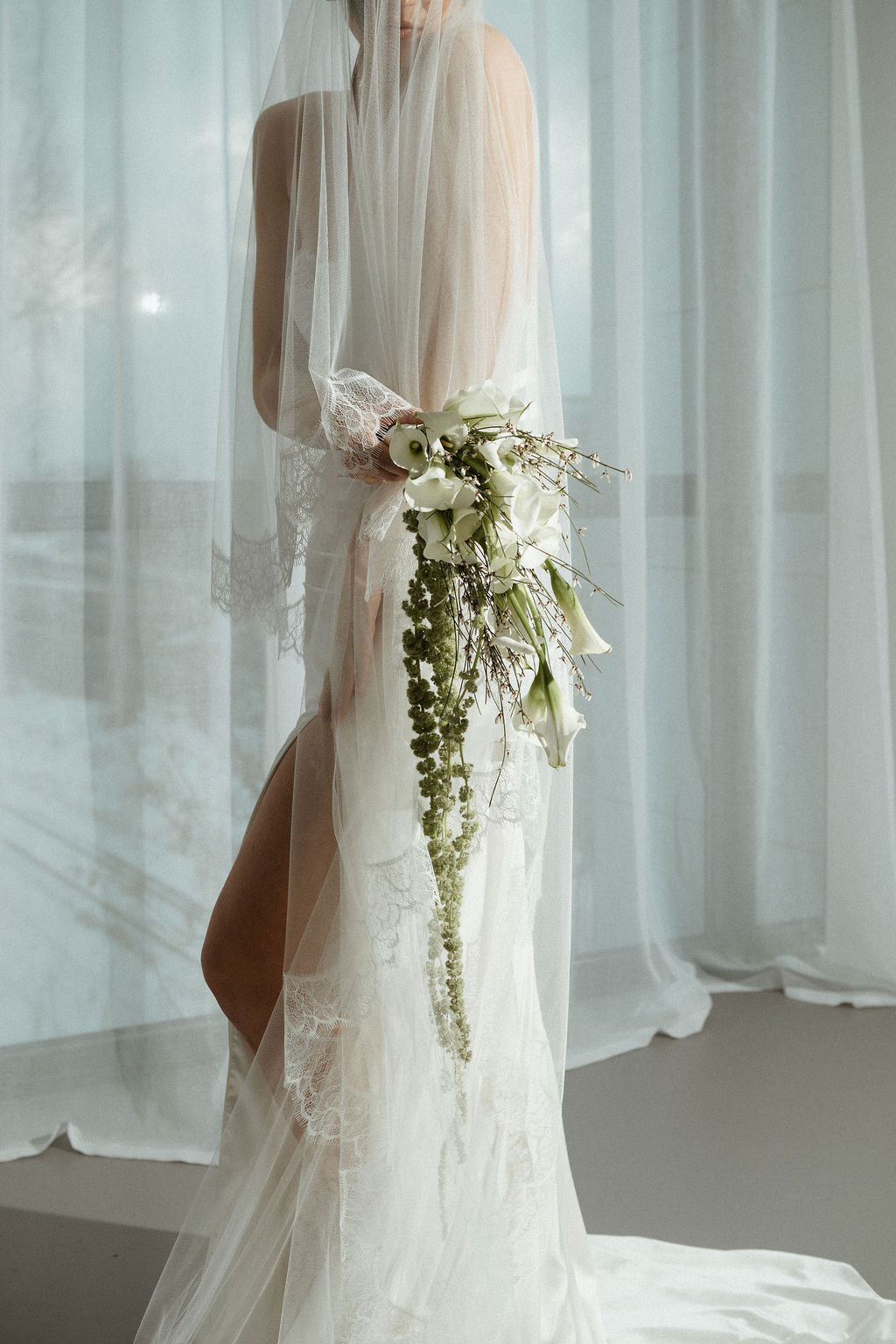 Person in a white wedding dress holding a floral bouquet with sheer curtains in the background