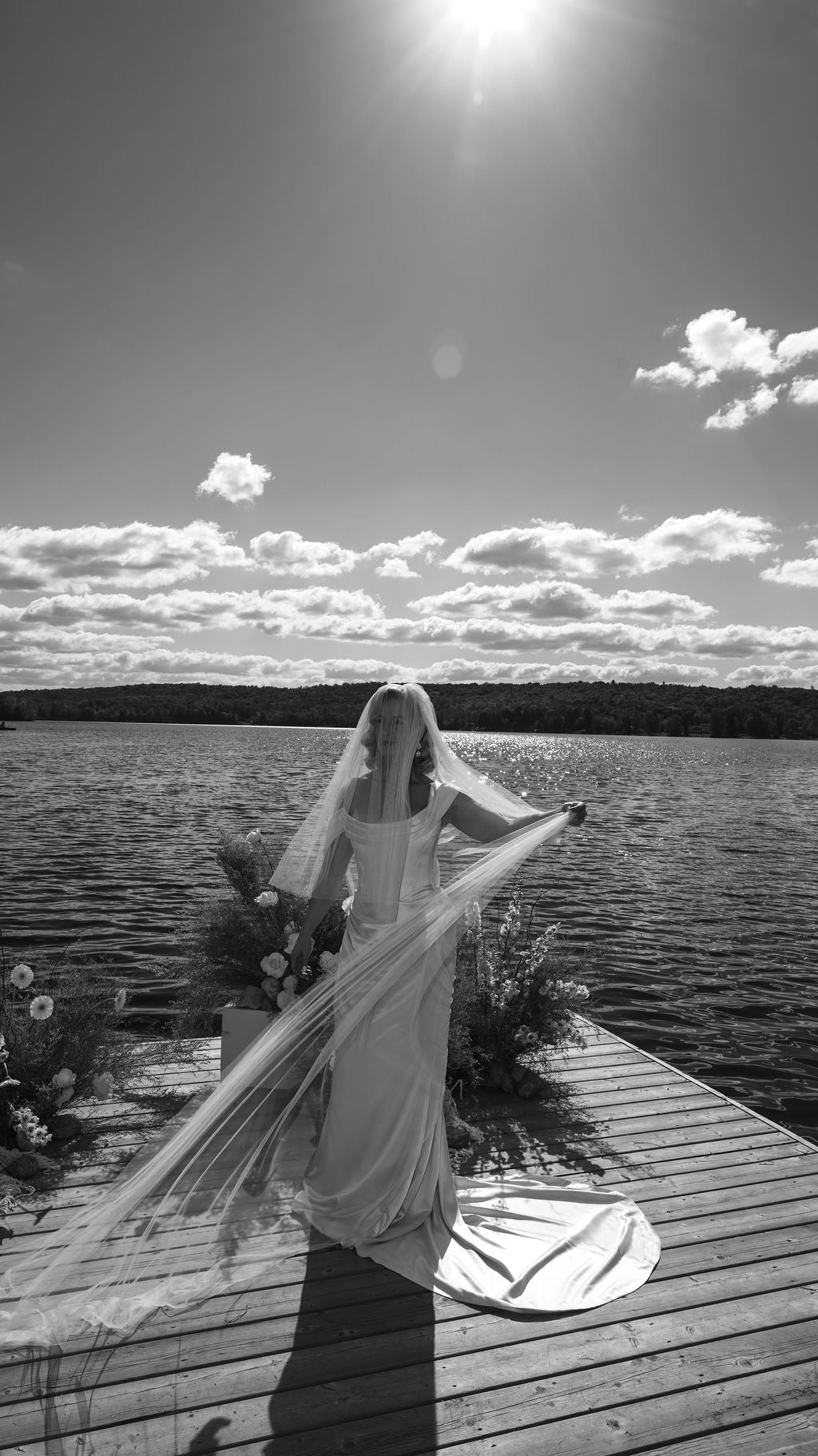 Woman in a wedding dress with a long veil standing on a dock by a lake.
