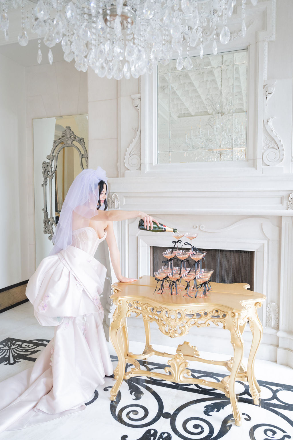 Woman in a white dress opening a bottle of champagne in a luxurious room with a chandelier and fireplace.