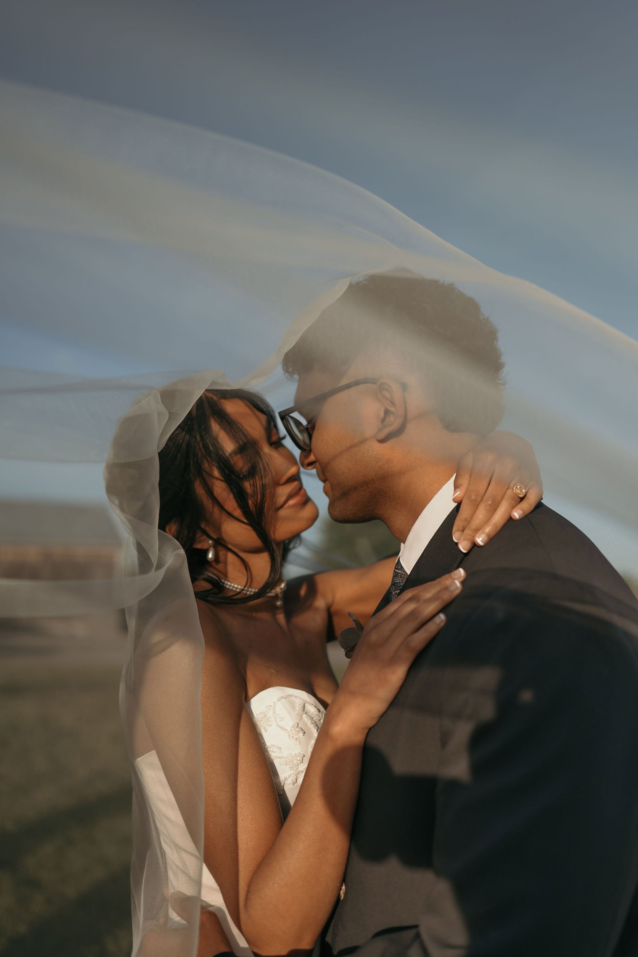 A bride in a white wedding dress and veil embracing and kissing her groom outdoors. The groom is wearing a dark suit and glasses, and a sheer veil is prominently visible against a soft blue sky background.