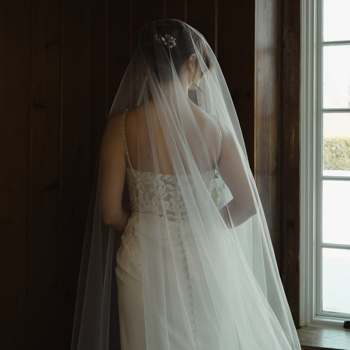 Bride in a white wedding dress with a long veil standing by a window.