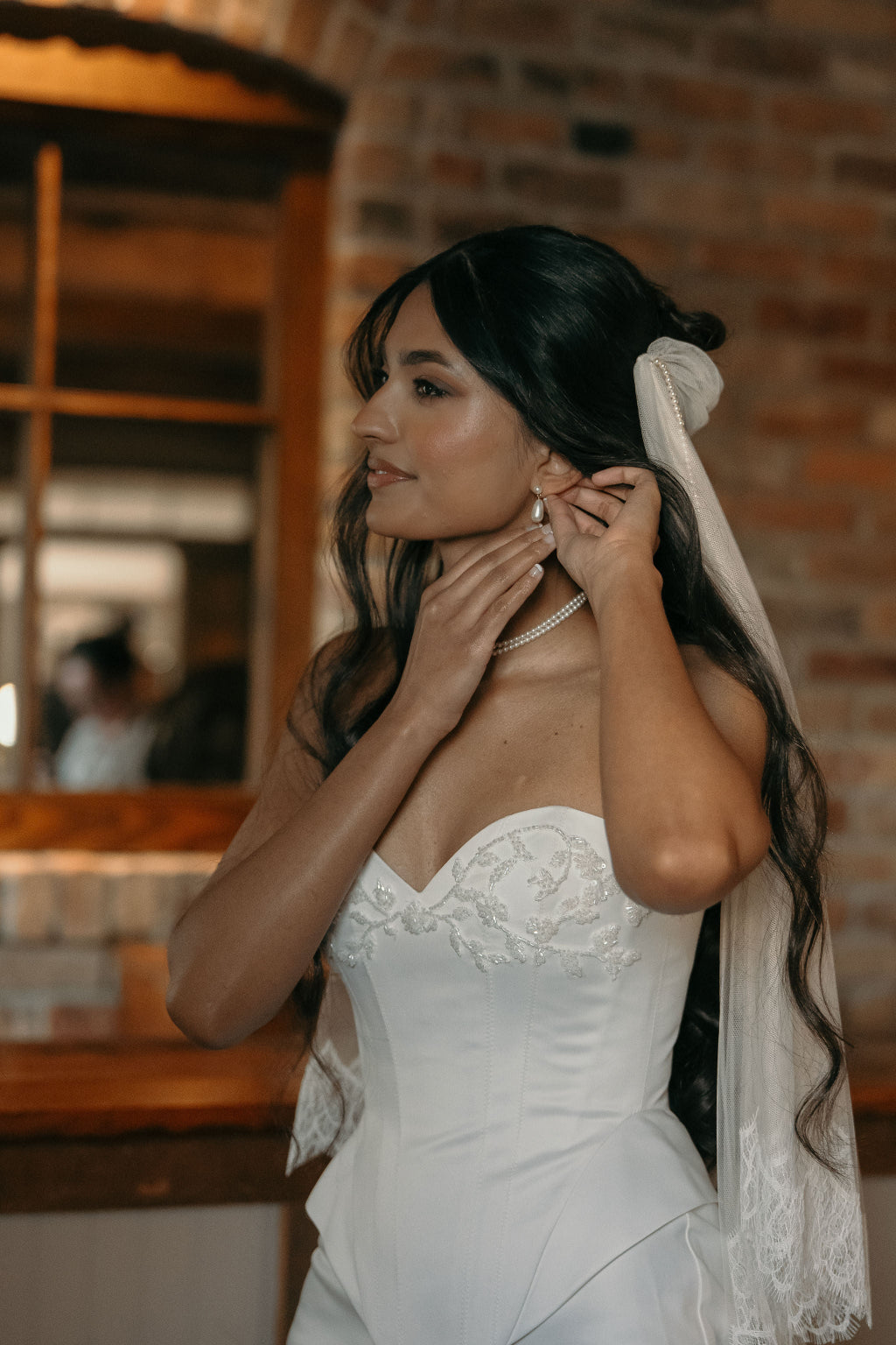 A woman in a white strapless lace wedding dress and veil adjusting her earring, standing indoors against a warm brick wall and wooden background.