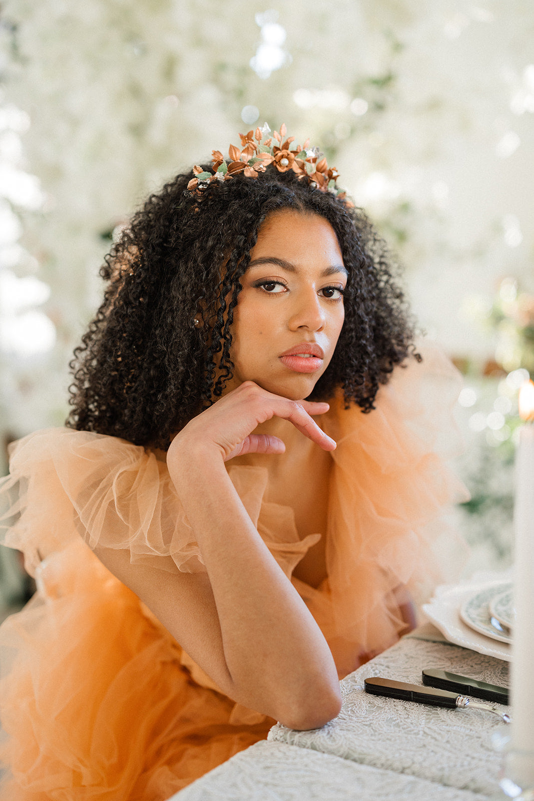 Woman in an orange dress with a tiara, sitting at a table with makeup items.