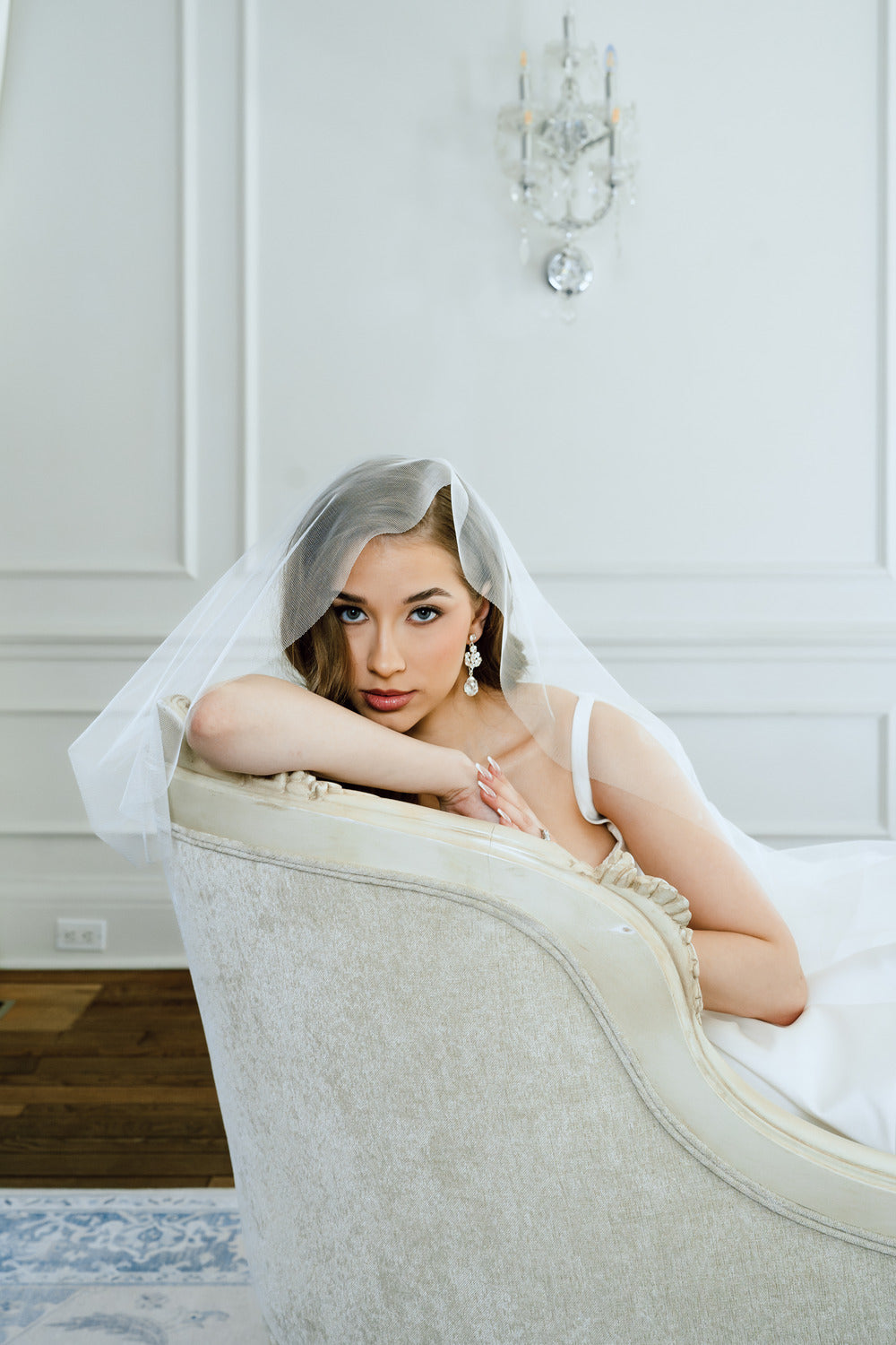 Woman in a white dress with a veil sitting on a white chair in a room with white walls.