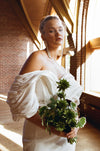 Woman in a white off-shoulder dress holding a green bouquet indoors.