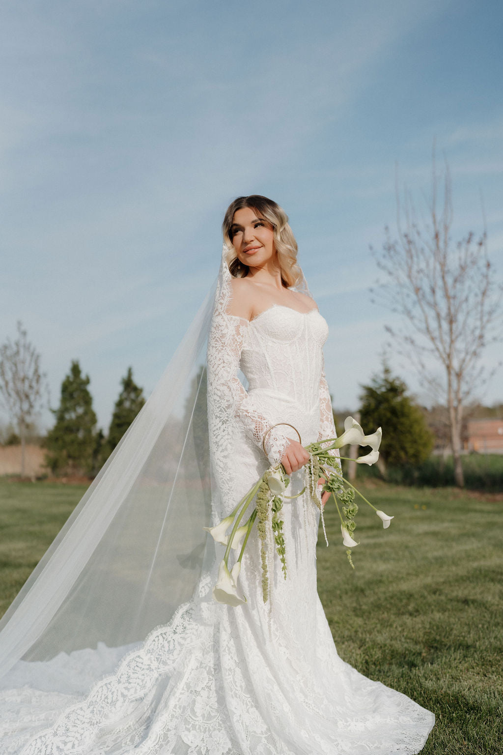 Woman in a white wedding dress holding a bouquet outdoors with trees and grass in the background