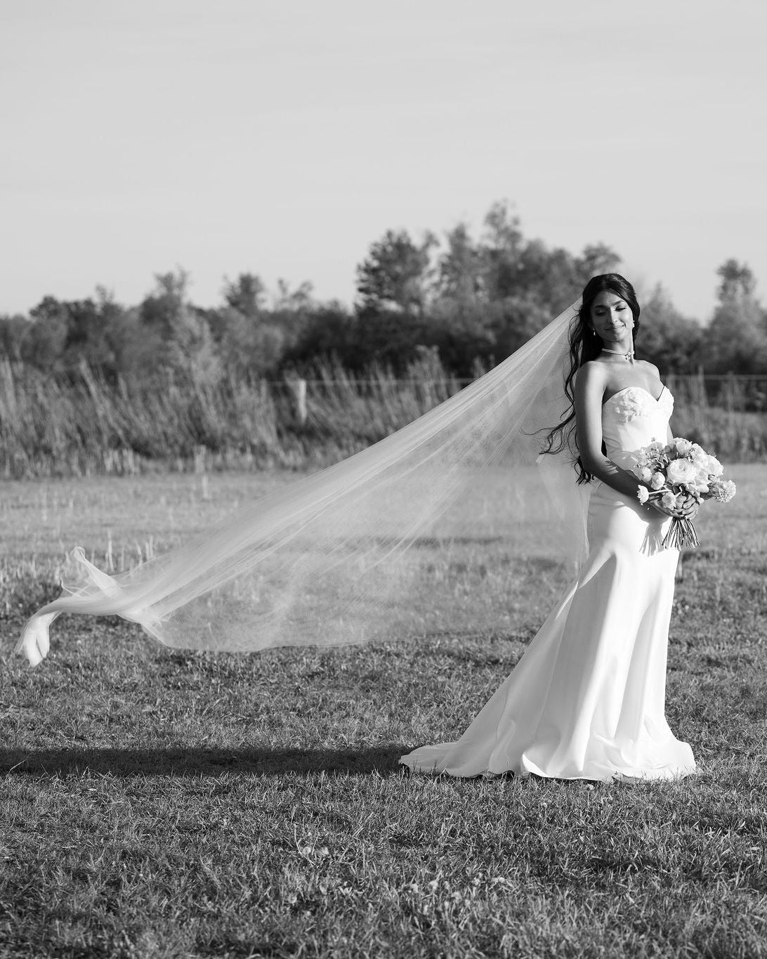 A black and white photograph of a bride standing in an open field, wearing a strapless wedding gown and holding a bouquet, with her long veil blowing in the wind.
