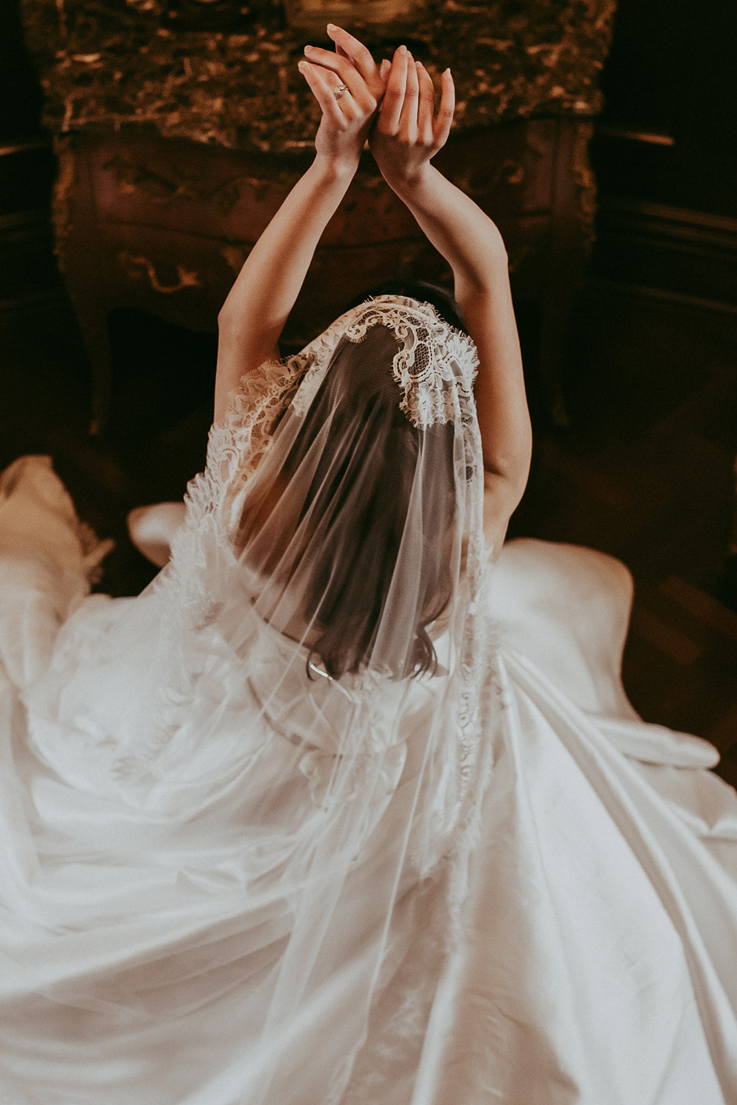 Person in a white wedding dress with a long veil, sitting with hands raised.