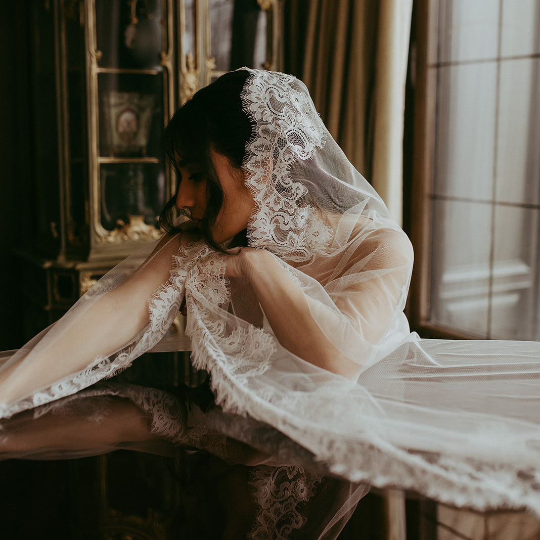 Woman in a lace veil sitting by a window in a room with ornate furniture.
