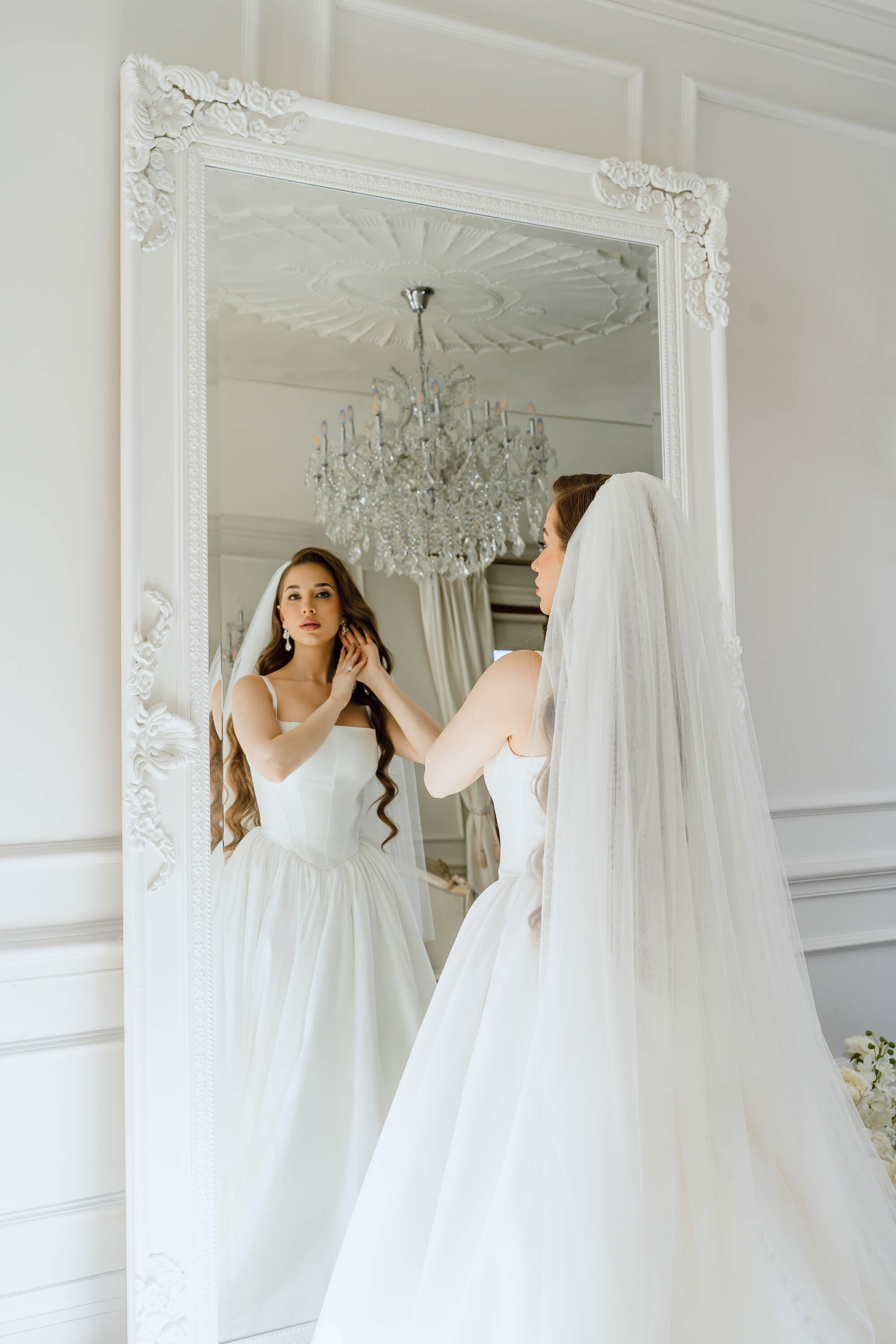 Bride in a white wedding dress standing in front of a large mirror, adjusting her veil.