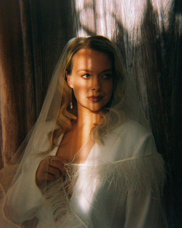 Woman in a white bridal outfit with a veil standing against a dark curtain.