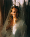 Woman in a white bridal outfit with a veil standing against a dark curtain.