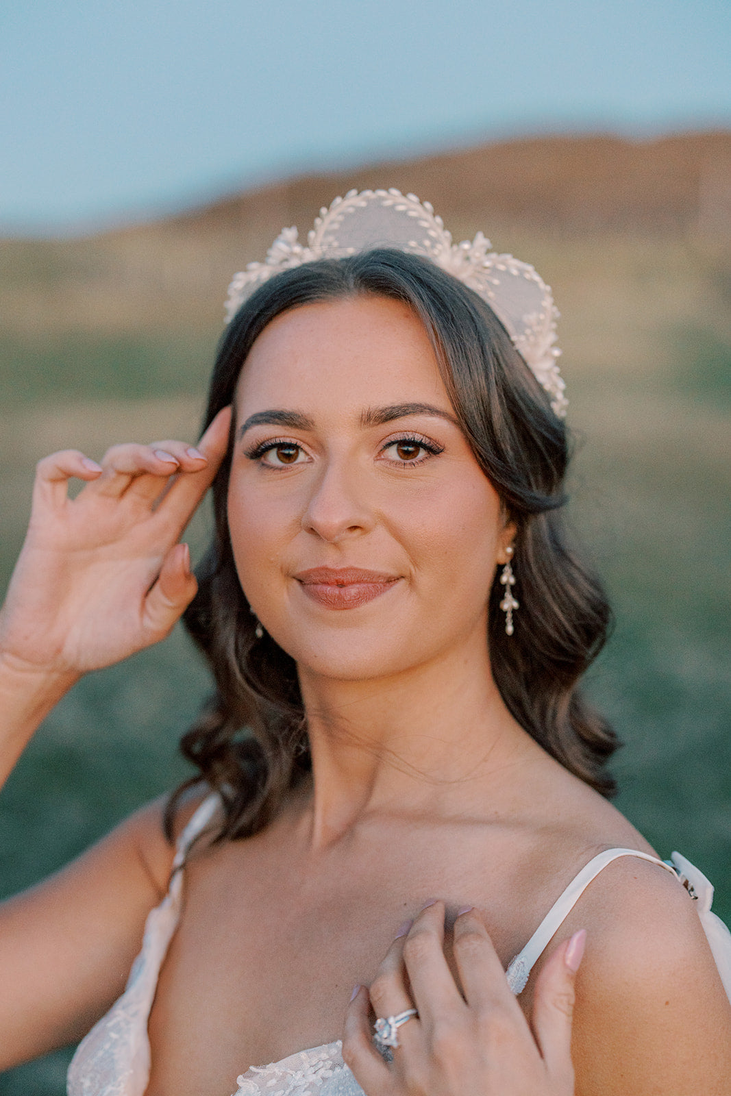 A woman outdoors wearing a white lace tiara and a white strap dress, with her hands near her face and a ring on one finger, set against a blurred natural background.