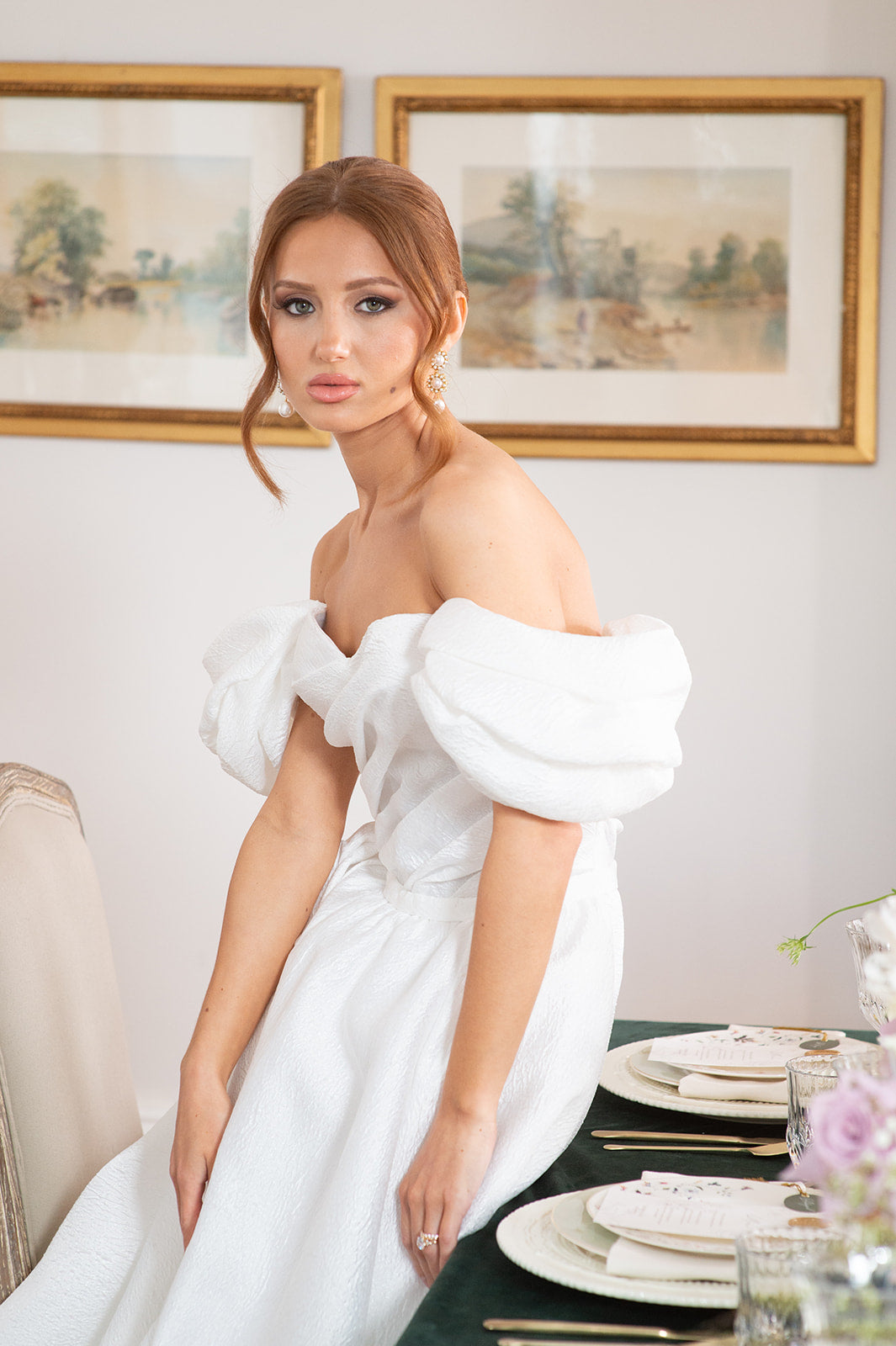 Woman in a white dress standing in a room with framed pictures on the wall.