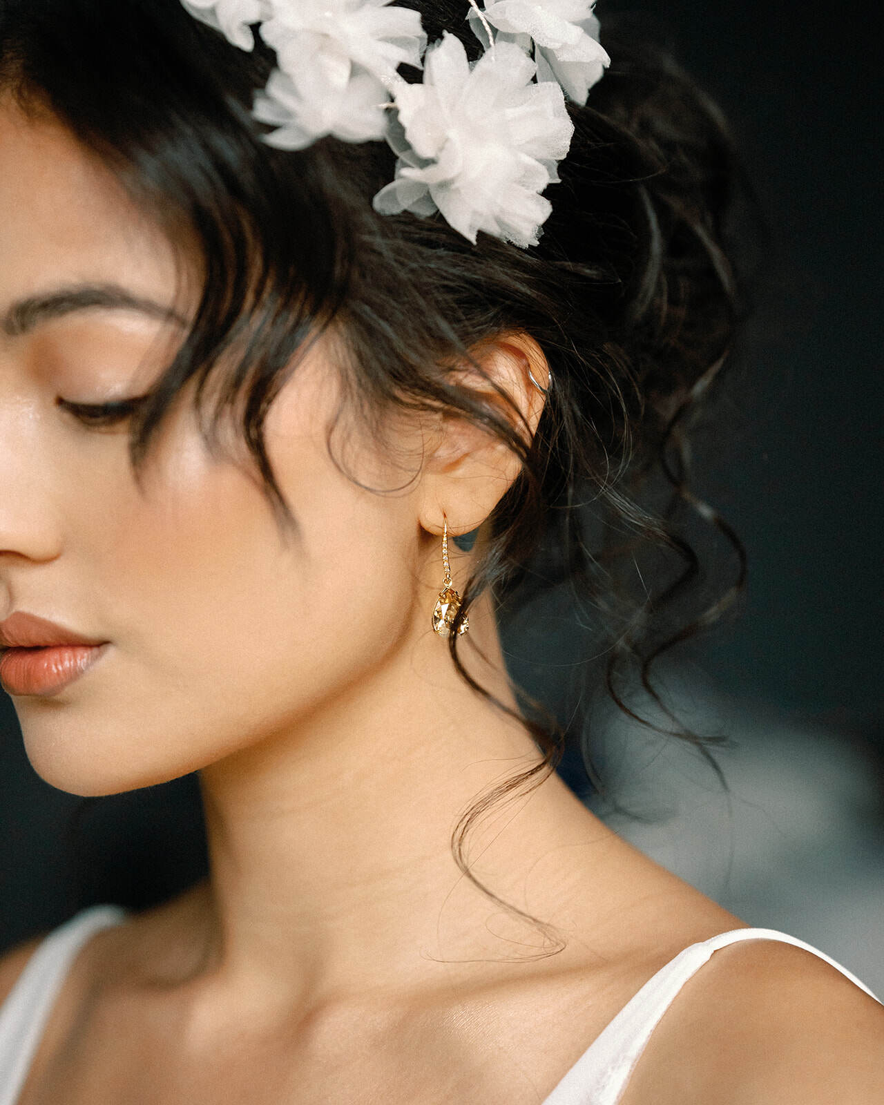 Woman wearing gold earrings with a white floral headband against a dark background