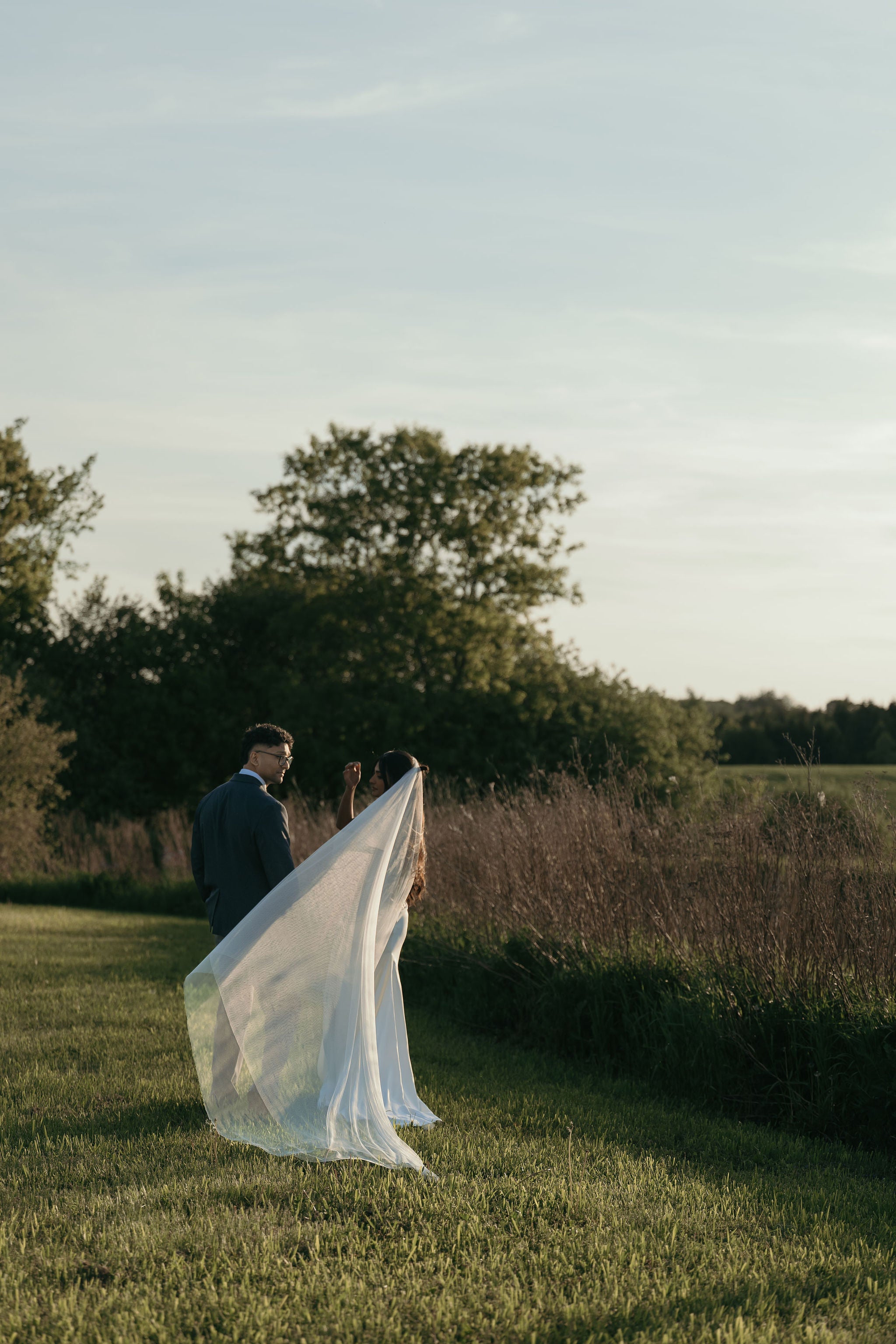 A bride in a flowing white wedding veil and a groom in a dark suit stand on a grassy field at sunset, with trees and tall grasses in the background.