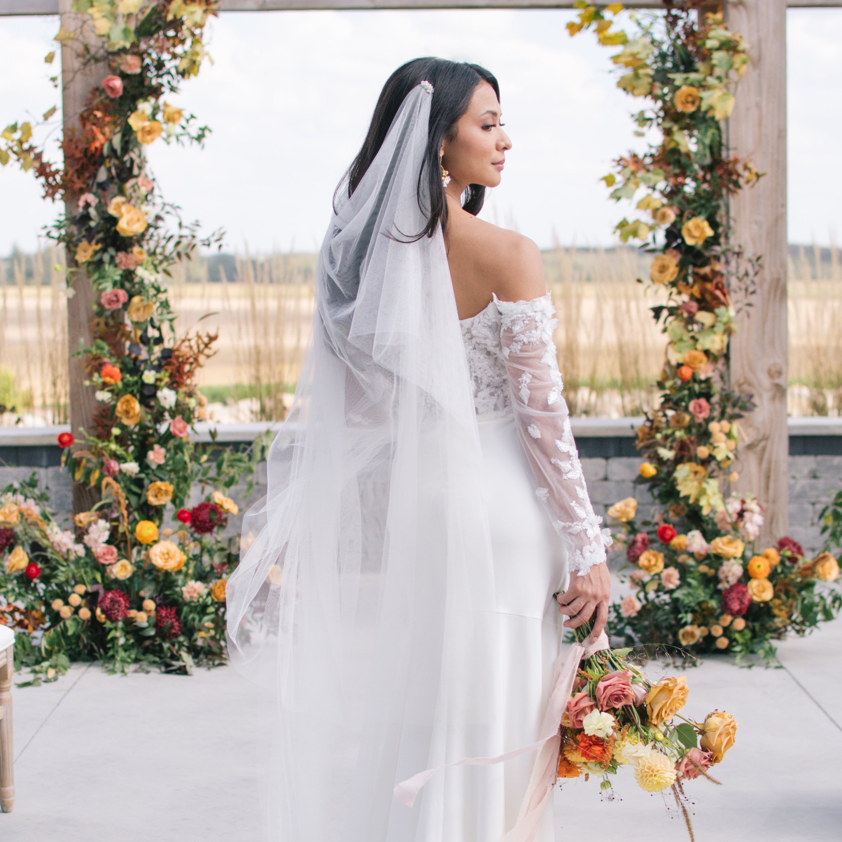 Bride in a white wedding dress with a long veil standing in front of floral arches.