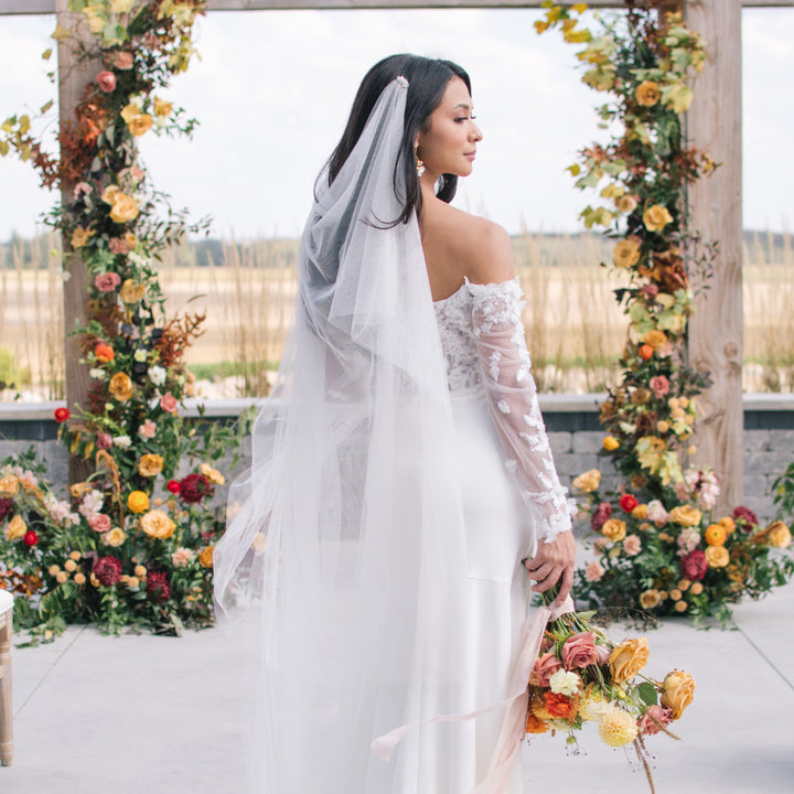 Bride in a white wedding dress with a long veil standing in front of floral arches.