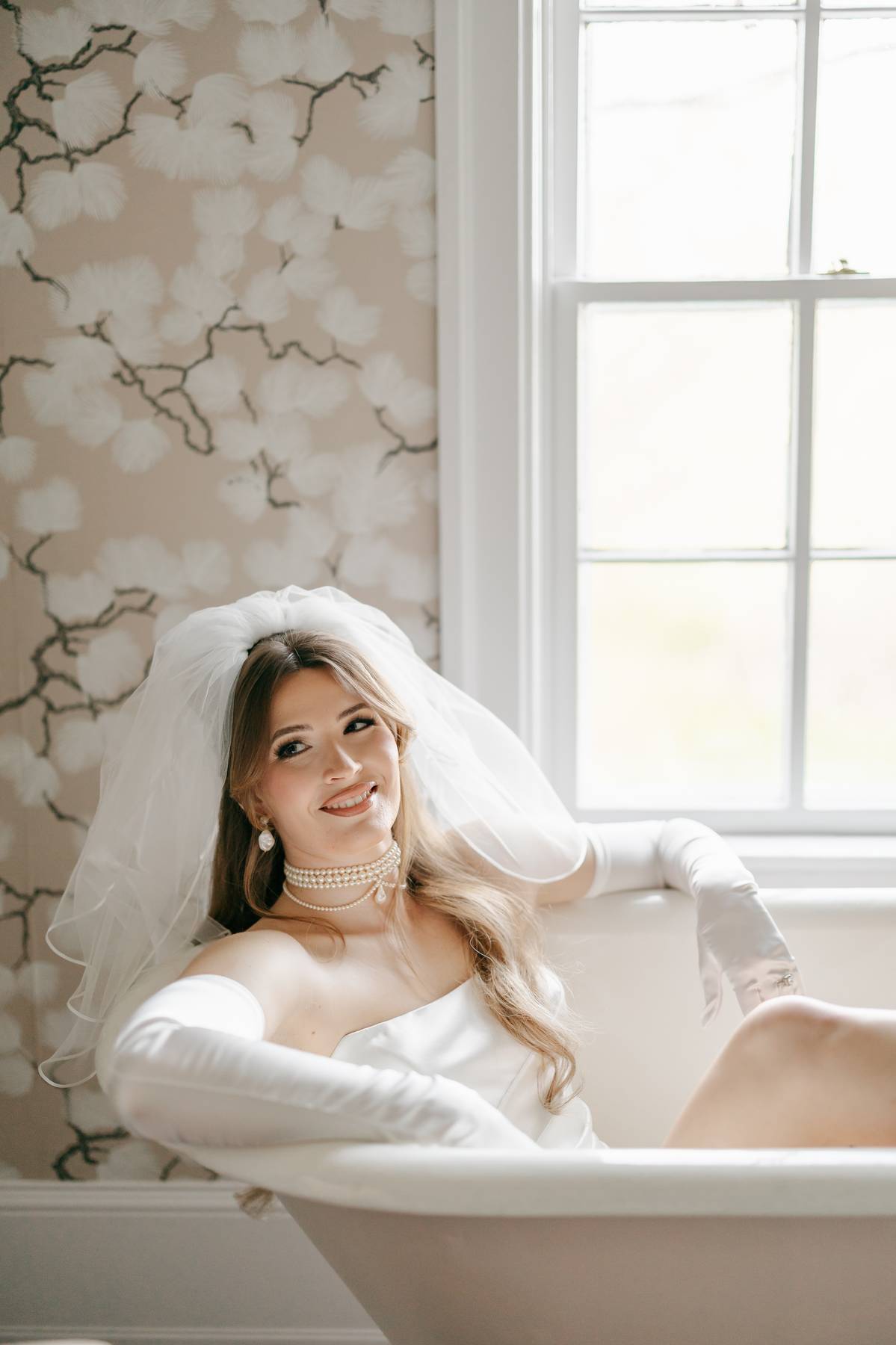 A smiling bride in a white wedding dress, veil, and long gloves sits in a white bathtub beside a window, with floral-patterned wallpaper in the background.