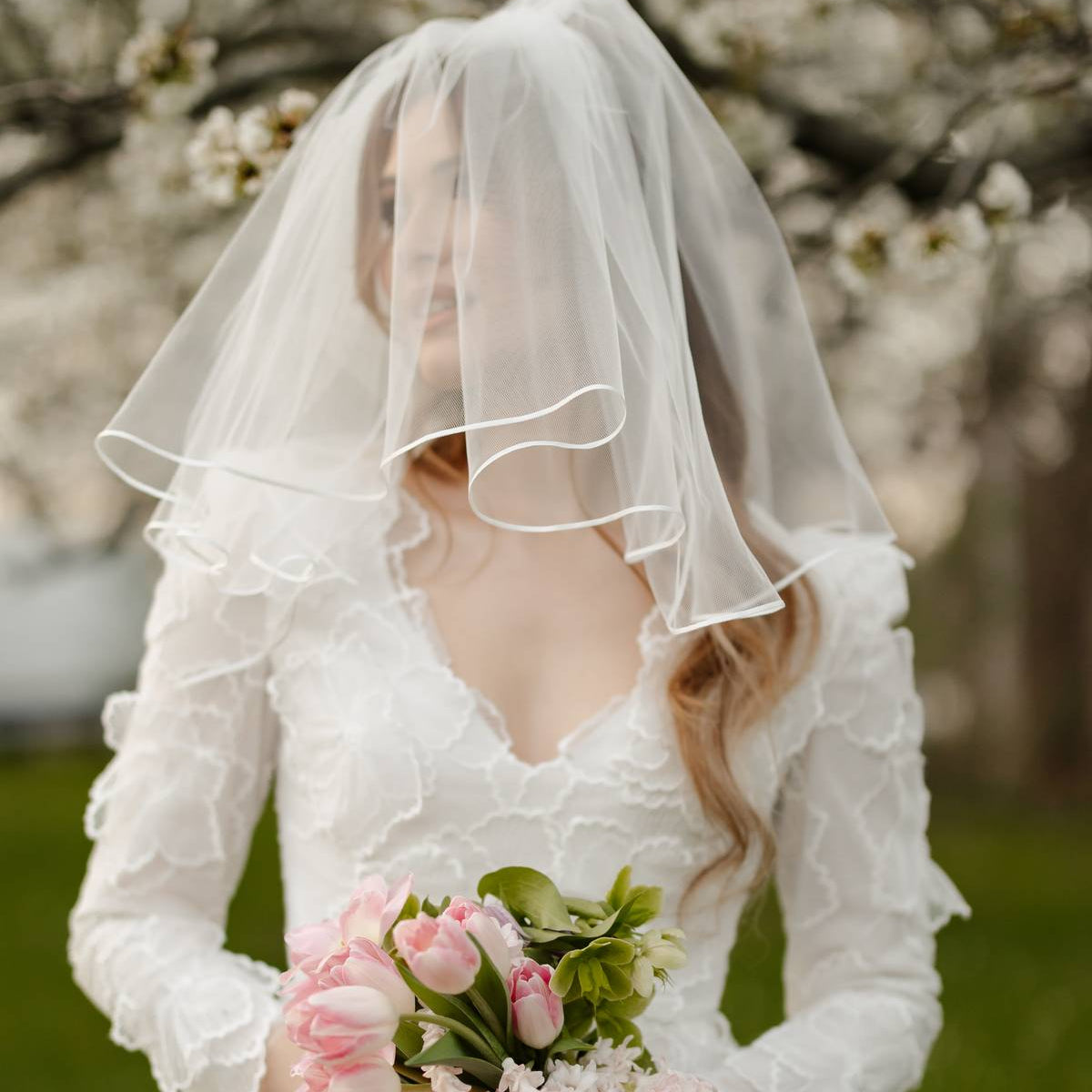 A bride in a white lace wedding dress with a sheer veil holds a bouquet of pink tulips and pale pink flowers outdoors among green grass and flowering tree branches.