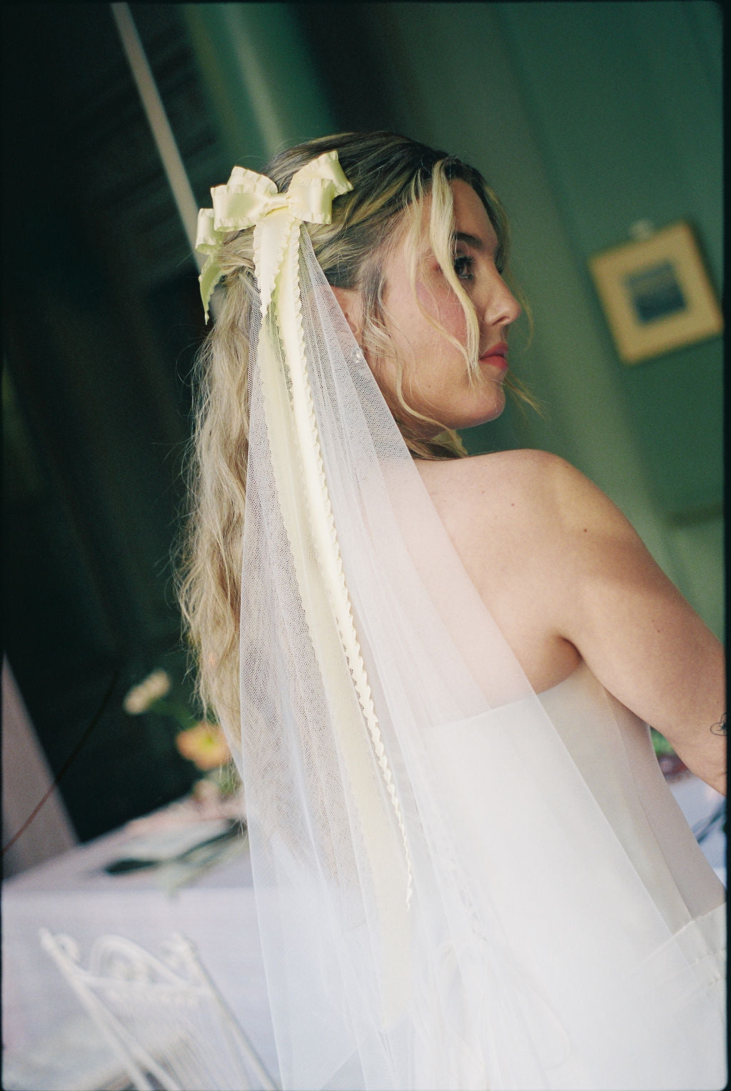 A woman in a white wedding dress seen from the back, wearing a long sheer veil with a pale yellow bow and ribbon in her hair, photographed indoors with a softly blurred green wall in the background.