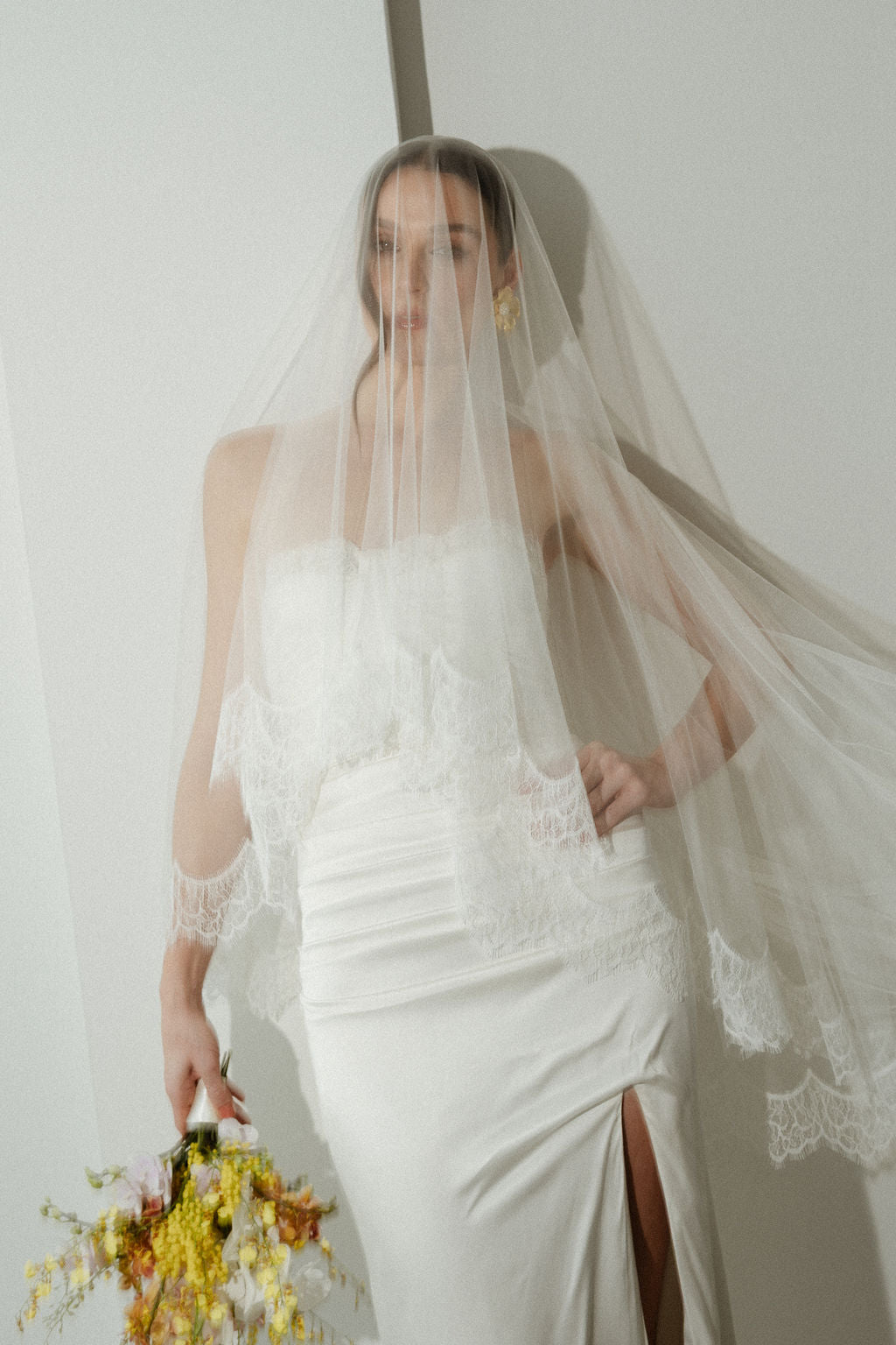 Woman in a white wedding dress with a veil holding flowers against a plain background