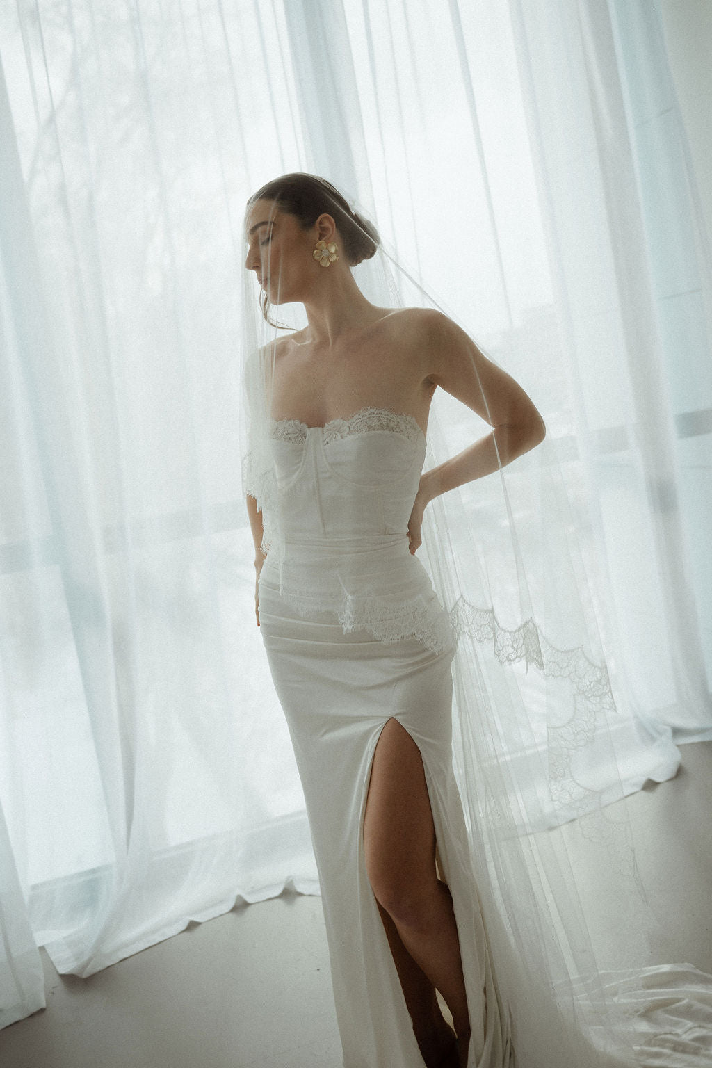 Woman wearing a white strapless gown with a slit in front, standing in front of sheer curtains.