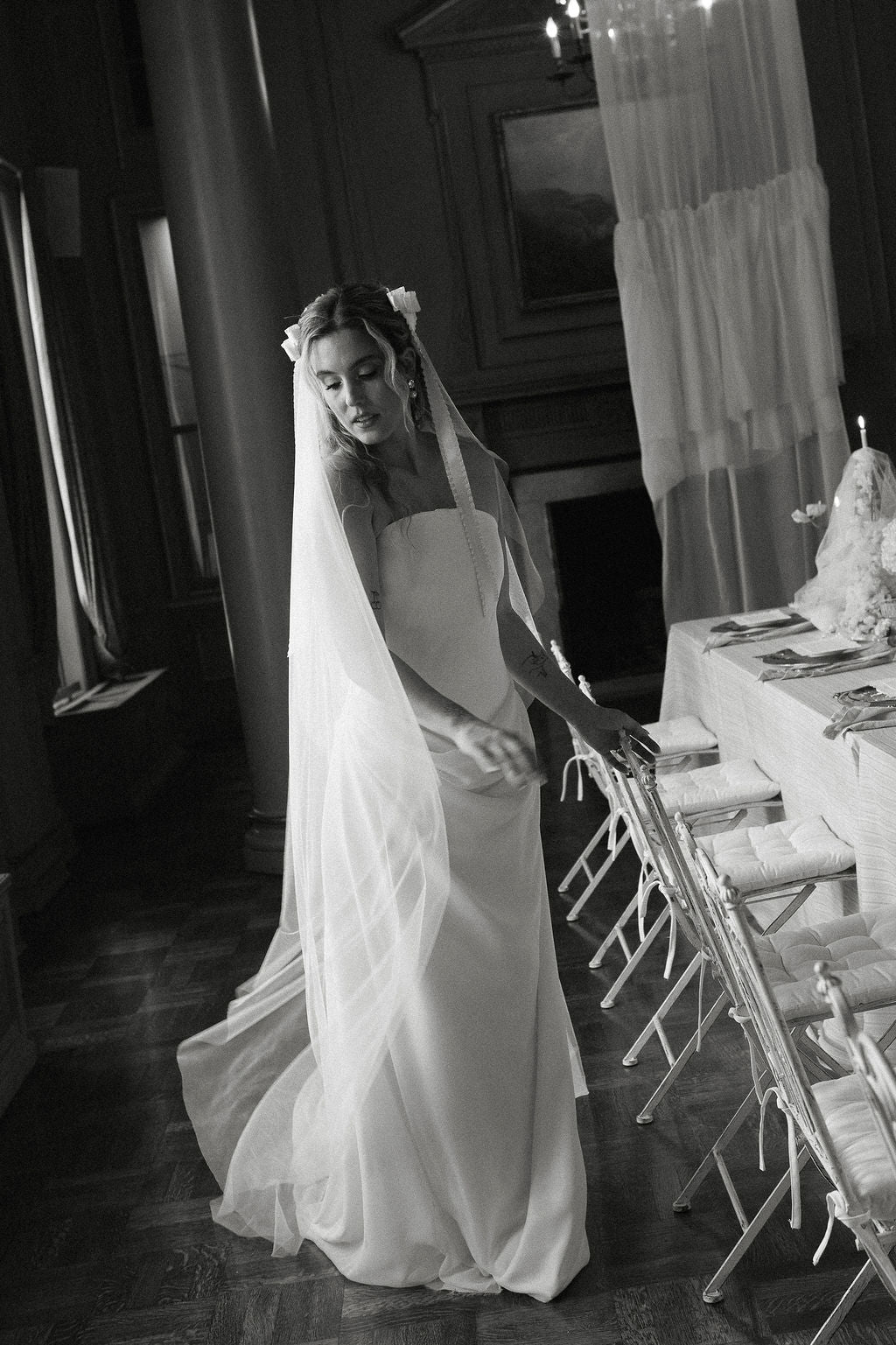 A black and white photograph of a bride in a strapless wedding gown and long veil, standing in a dimly lit hall next to a table with chairs.
