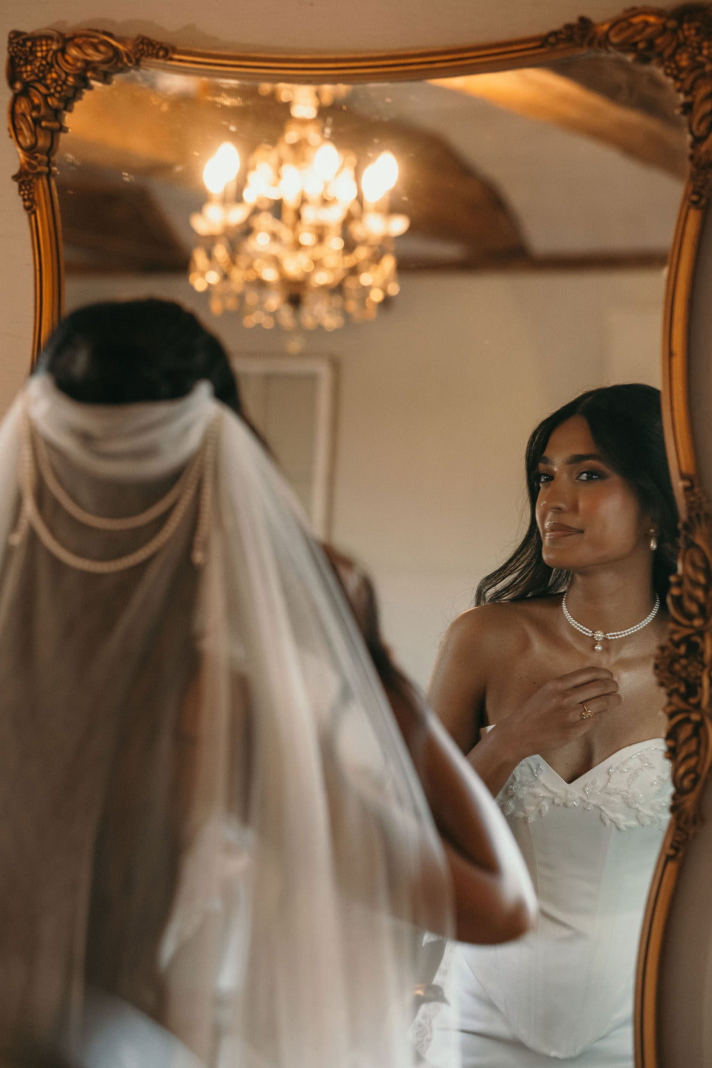 Woman adjusting her necklace in front of a mirror with a chandelier in the background