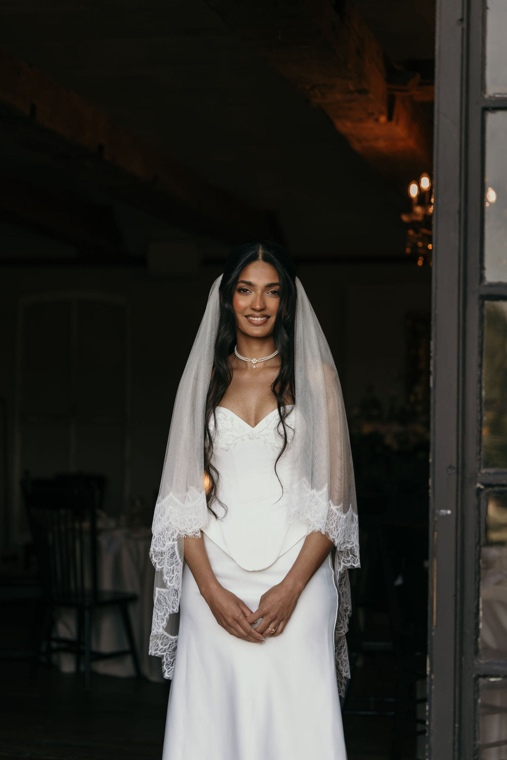 Woman in a white wedding dress with a veil standing in a dimly lit room.
