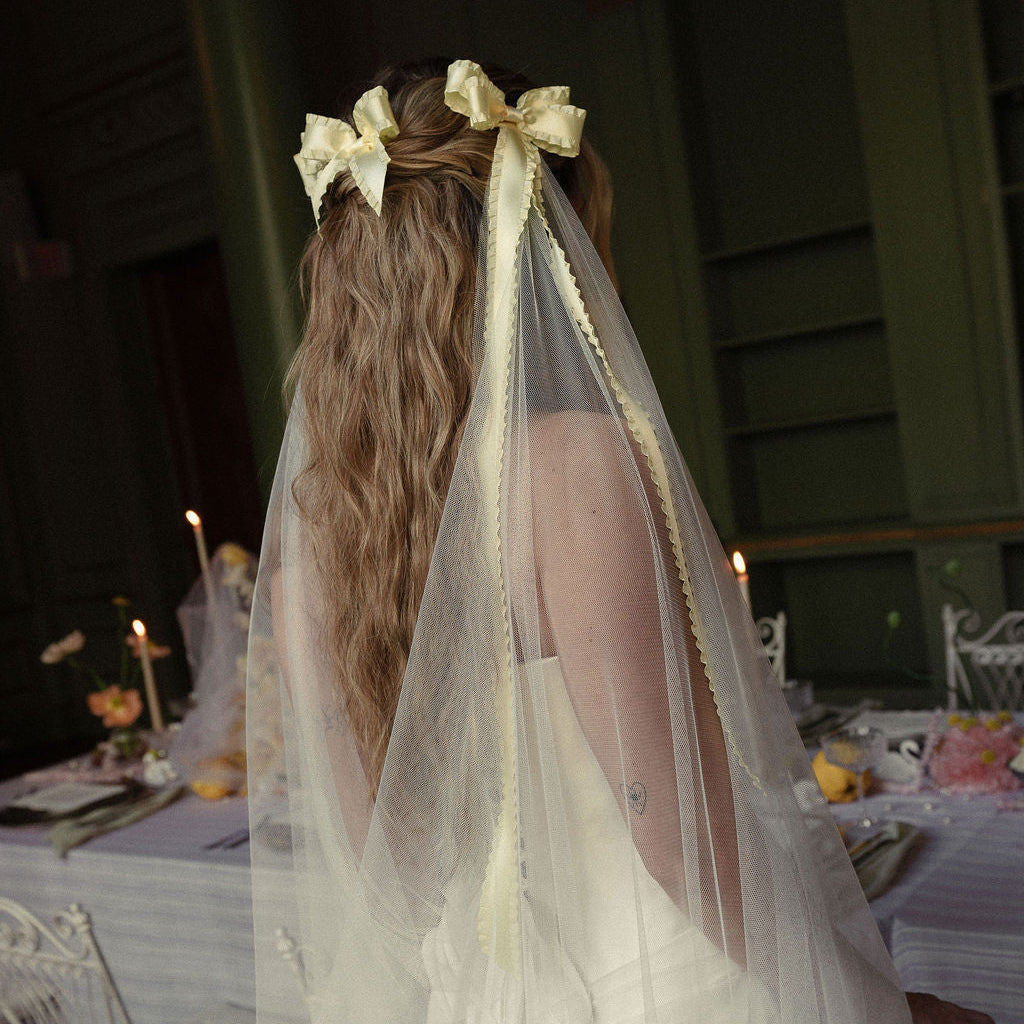 A woman seen from behind wearing a white wedding dress and a sheer veil with cream ribbon bows in her hair, standing in a dimly lit indoor event space with tables and candles in the background.