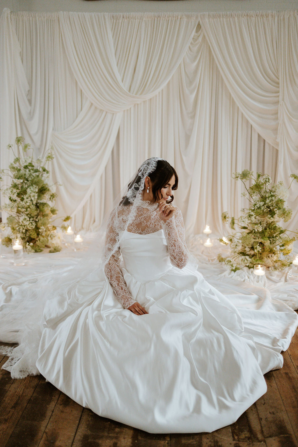 Woman in a white wedding dress sitting in front of draped curtains with floral arrangements.