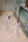 Two women in wedding dresses holding hands in a gallery setting with art on the walls.