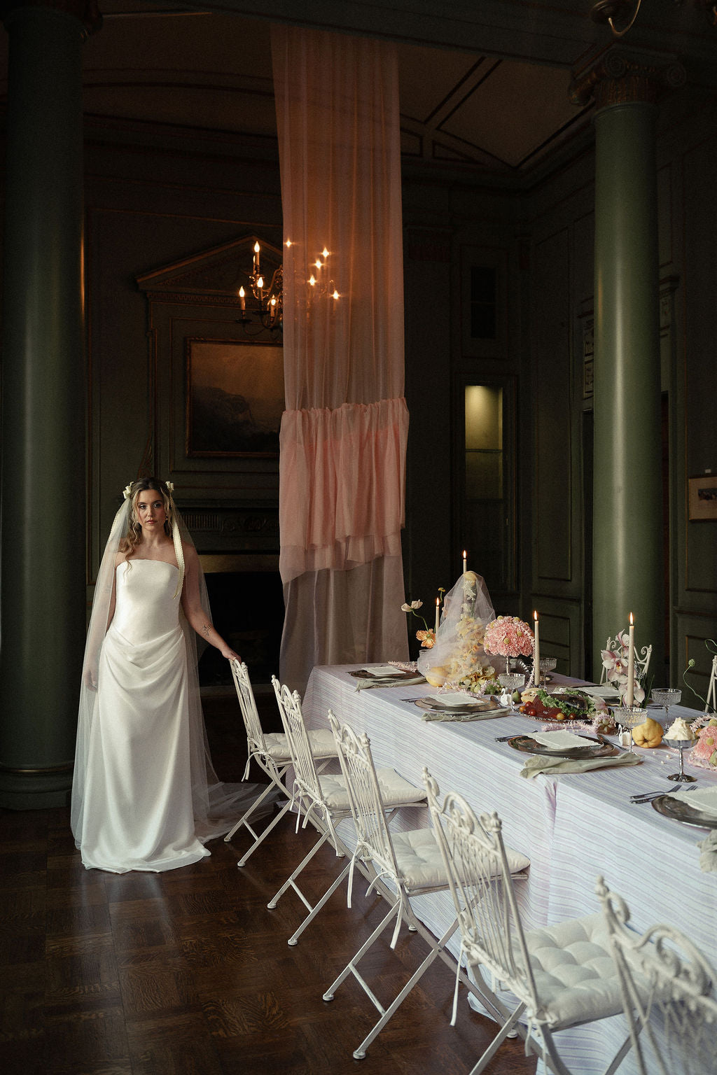 A bride in a white sleeveless wedding gown and veil walks beside a long wedding reception table set with white chairs, floral centerpieces, and a draped pink fabric in a dark, elegant hall.