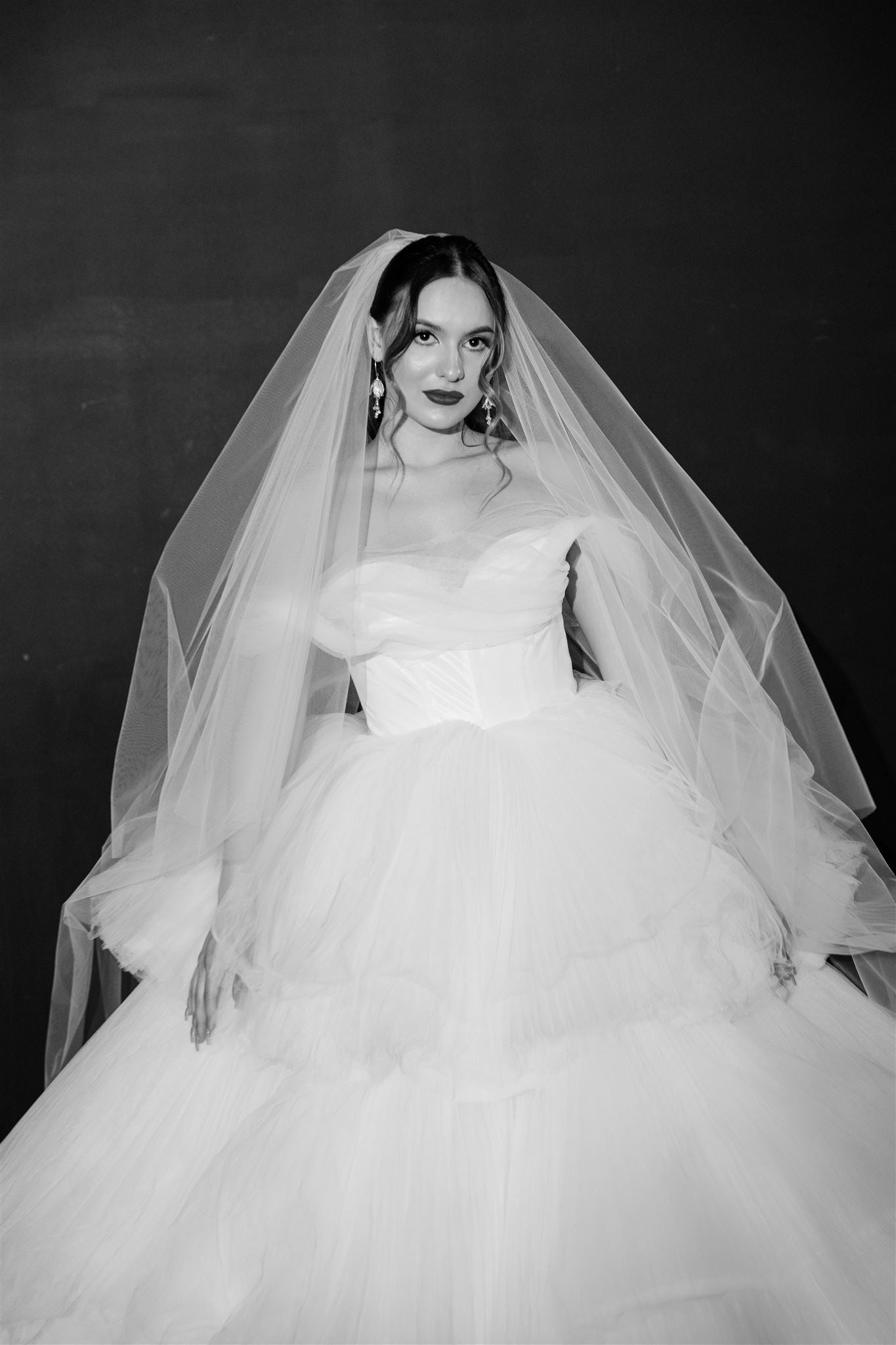 Black and white photo of a bride in a wedding dress with a long veil against a dark background
